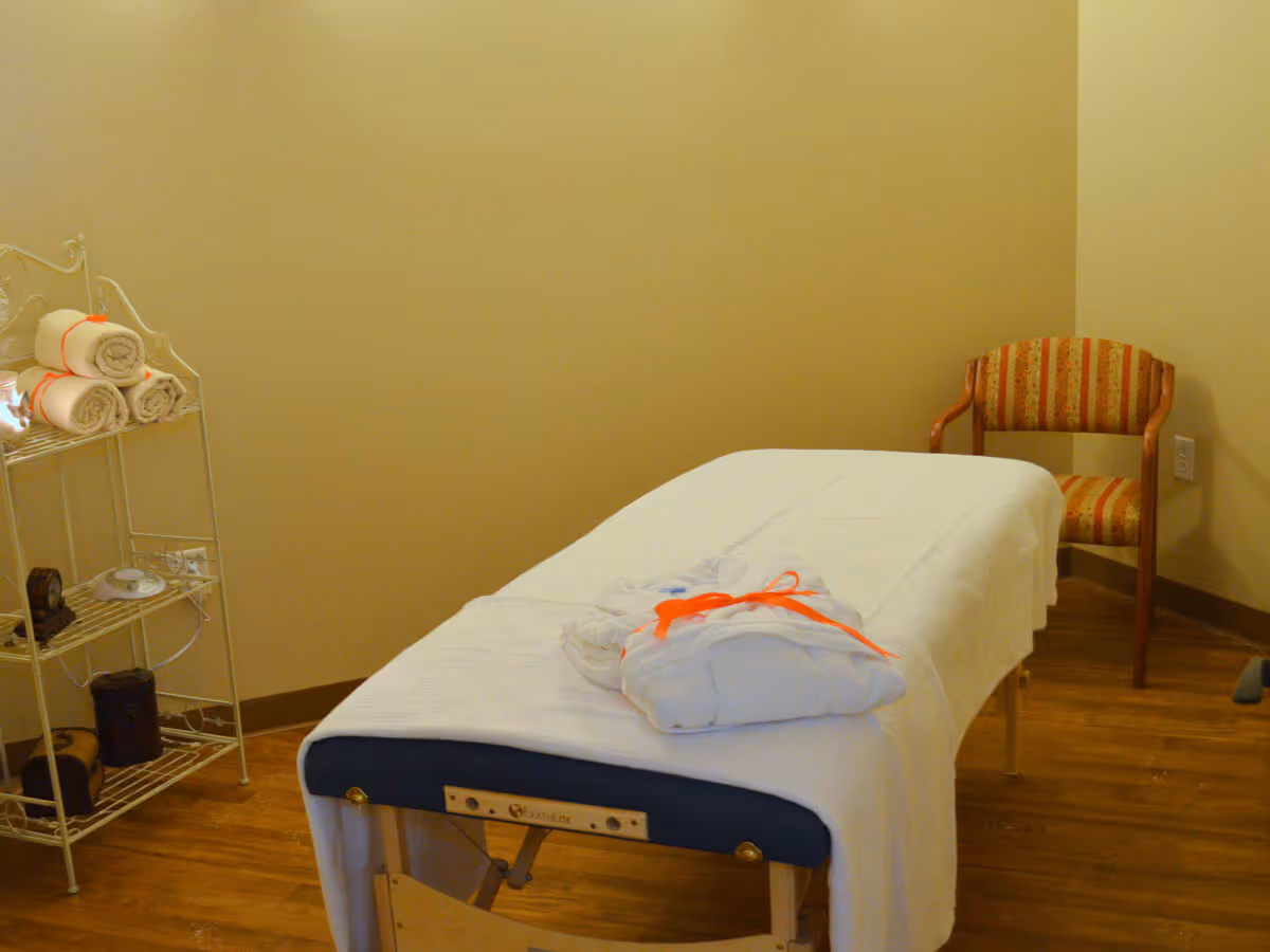 A small treatment room with a massage table draped in white linens, a shelf of rolled towels, and a striped chair.