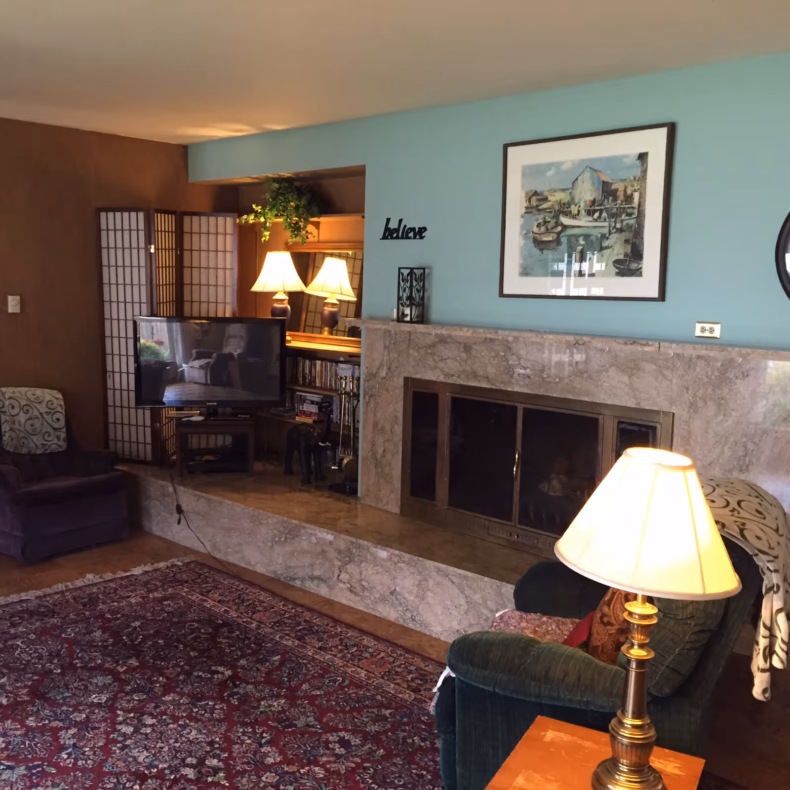 Cozy living room with a large patterned rug, a green armchair with a lamp on a wooden side table, a purple armchair, a TV on a stand, a decorative folding screen, and a marble fireplace with a framed painting above it. The wall behind the fireplace is painted light blue and has the word 'believe' displayed on it.