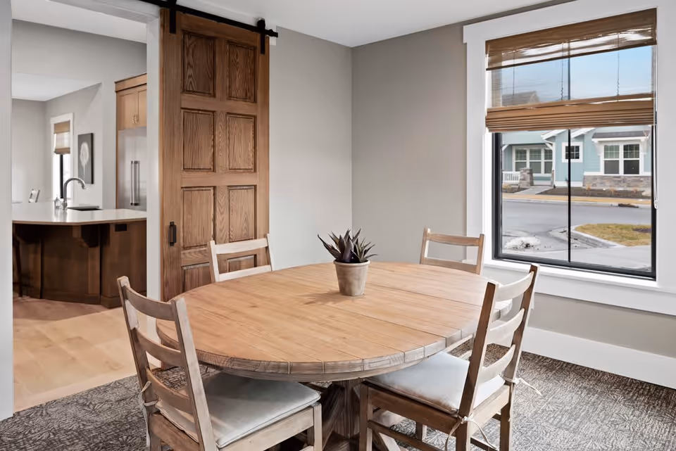 A dining area with a round wooden table surrounded by four wooden chairs with cushions. A small potted plant is placed in the center of the table. There is a large window with a brown blind partially open, showing a view of a street and houses outside. To the left, a sliding wooden barn door is partially open, revealing a kitchen area with a countertop and a sink.