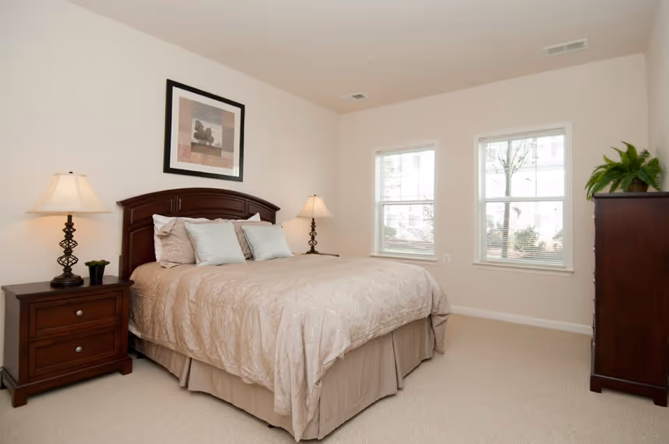 A neatly made bed with beige bedding and multiple pillows in a bedroom. There are two bedside tables with lamps on each side of the bed. A framed picture hangs above the headboard. Two windows with blinds allow natural light into the room. A dark wooden dresser with a green plant on top is positioned against the right wall.