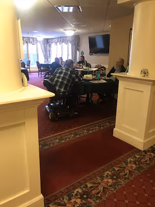 A group of elderly residents seated around tables in a carpeted communal dining/activity room with a wall-mounted TV and large windows.