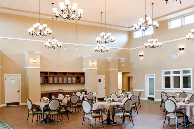 A spacious dining room with multiple round tables covered with beige tablecloths, each surrounded by cushioned chairs. The room features high ceilings with several elegant chandeliers hanging down, beige walls, and large windows near the ceiling allowing natural light to enter. There are cabinets and a countertop along one wall, and doors leading to other areas.