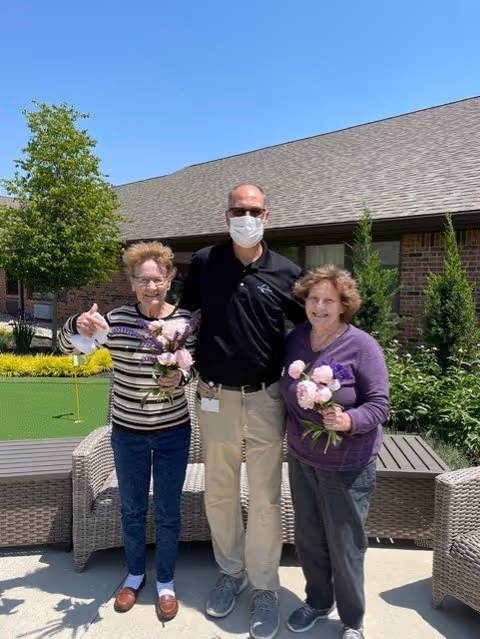 Two elderly women holding flowers stand outside with a man wearing a face mask and sunglasses between them. They are smiling and posing for the photo in a sunny outdoor area with wicker chairs, green plants, and a building with a shingled roof in the background.