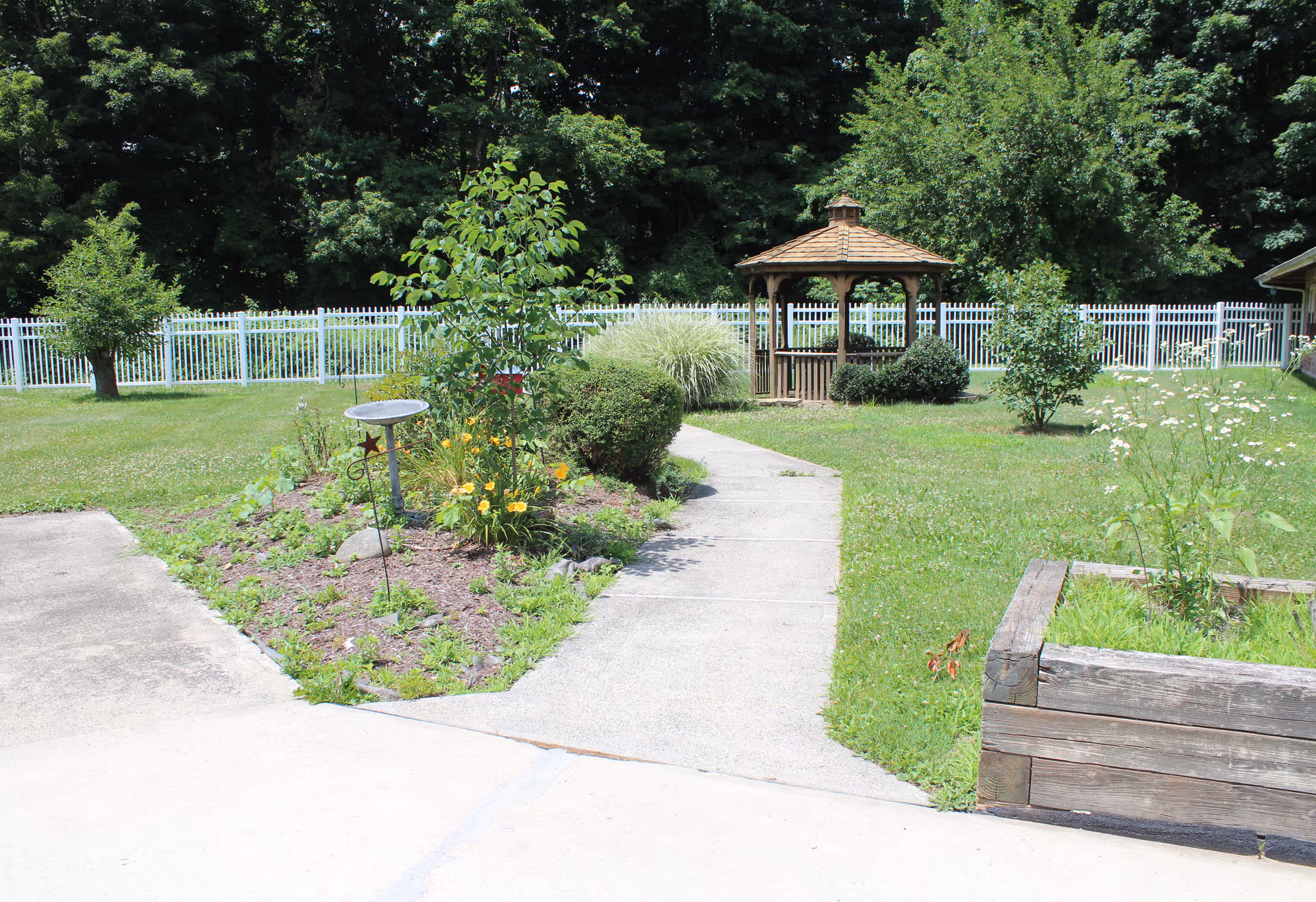 A garden area with a concrete pathway leading to a wooden gazebo surrounded by green grass, bushes, and trees. There is a white fence in the background and a raised wooden planter box with plants on the right side.