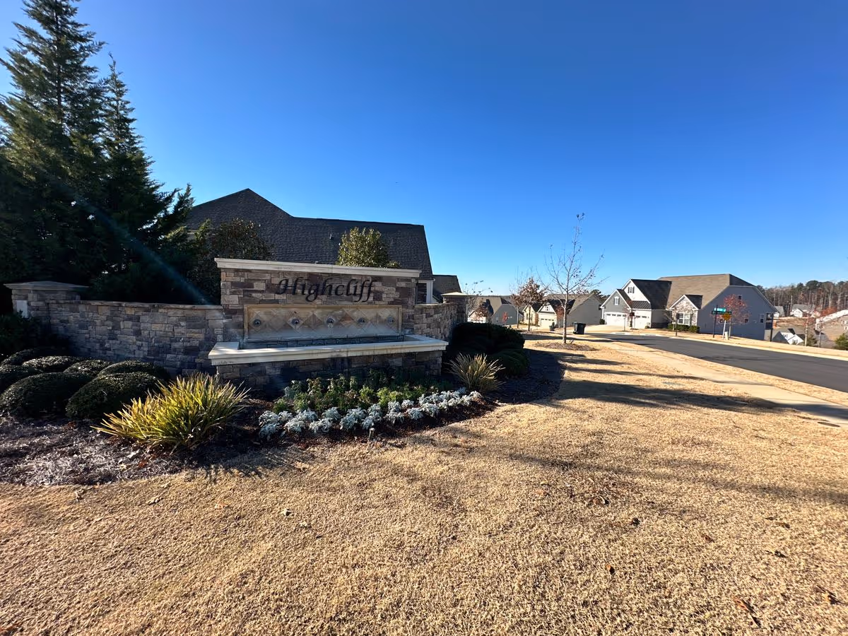 Stone entrance sign reading "Highcliff" marking a landscaped neighborhood entrance with houses and a clear blue sky.