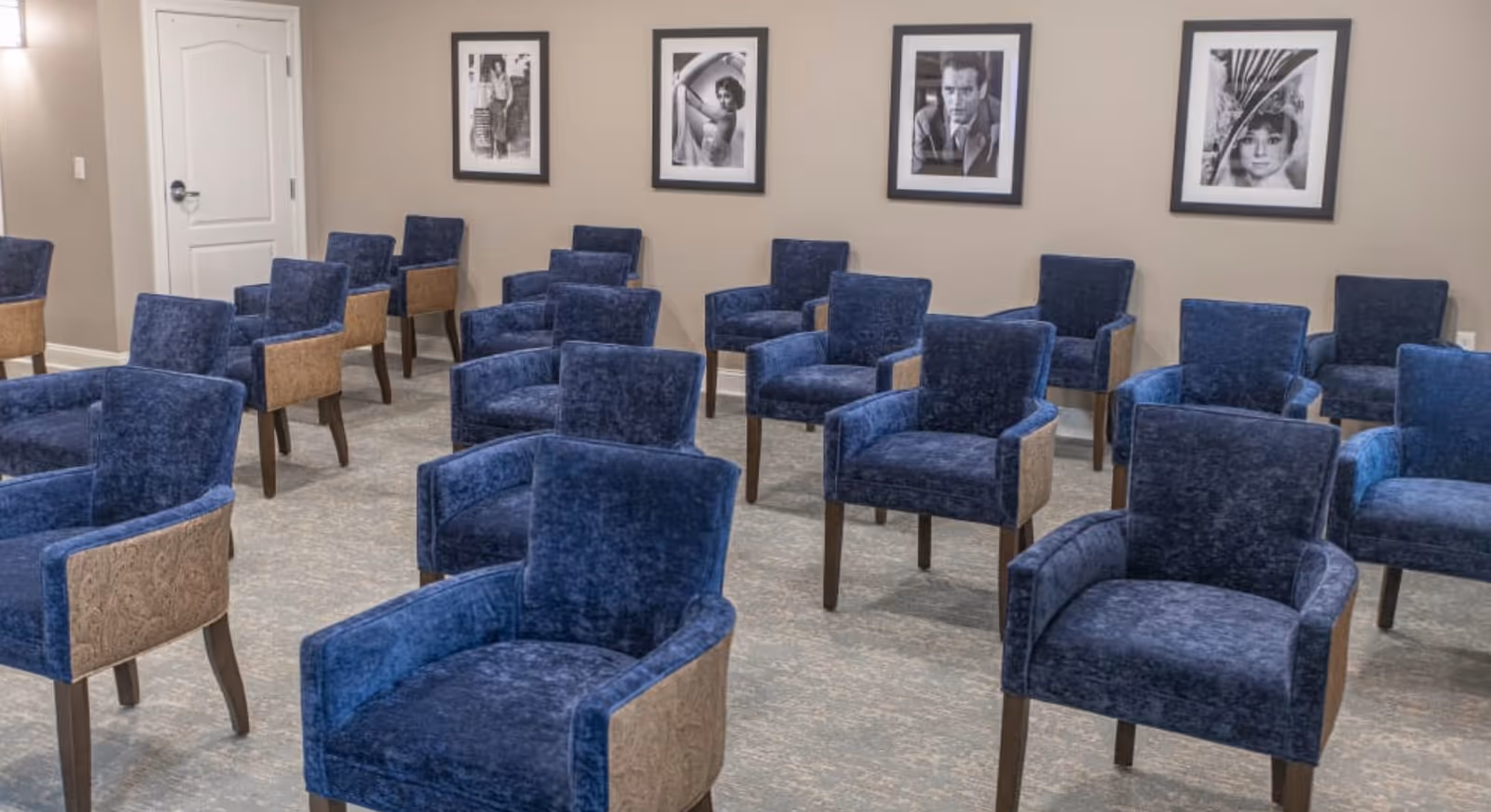 A room with multiple blue upholstered chairs arranged in rows facing the same direction. The walls are beige and decorated with four black and white framed photographs. There is a white door on the left side of the room.