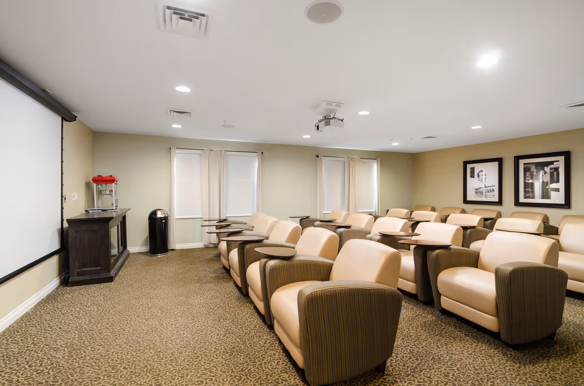 A small theater room with multiple rows of beige and brown cushioned armchairs, each equipped with a small attached table. The room has a large projector screen on one wall, a popcorn machine on a cabinet, and framed black-and-white photos on the opposite wall. The carpet is patterned, and the room is softly lit with recessed ceiling lights.