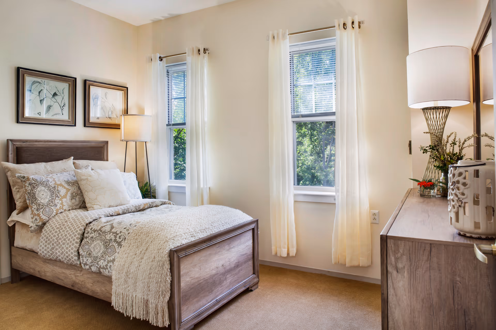 Sunlit bedroom with a wooden bed, patterned bedding, two windows with sheer curtains, a dresser, and lamps.