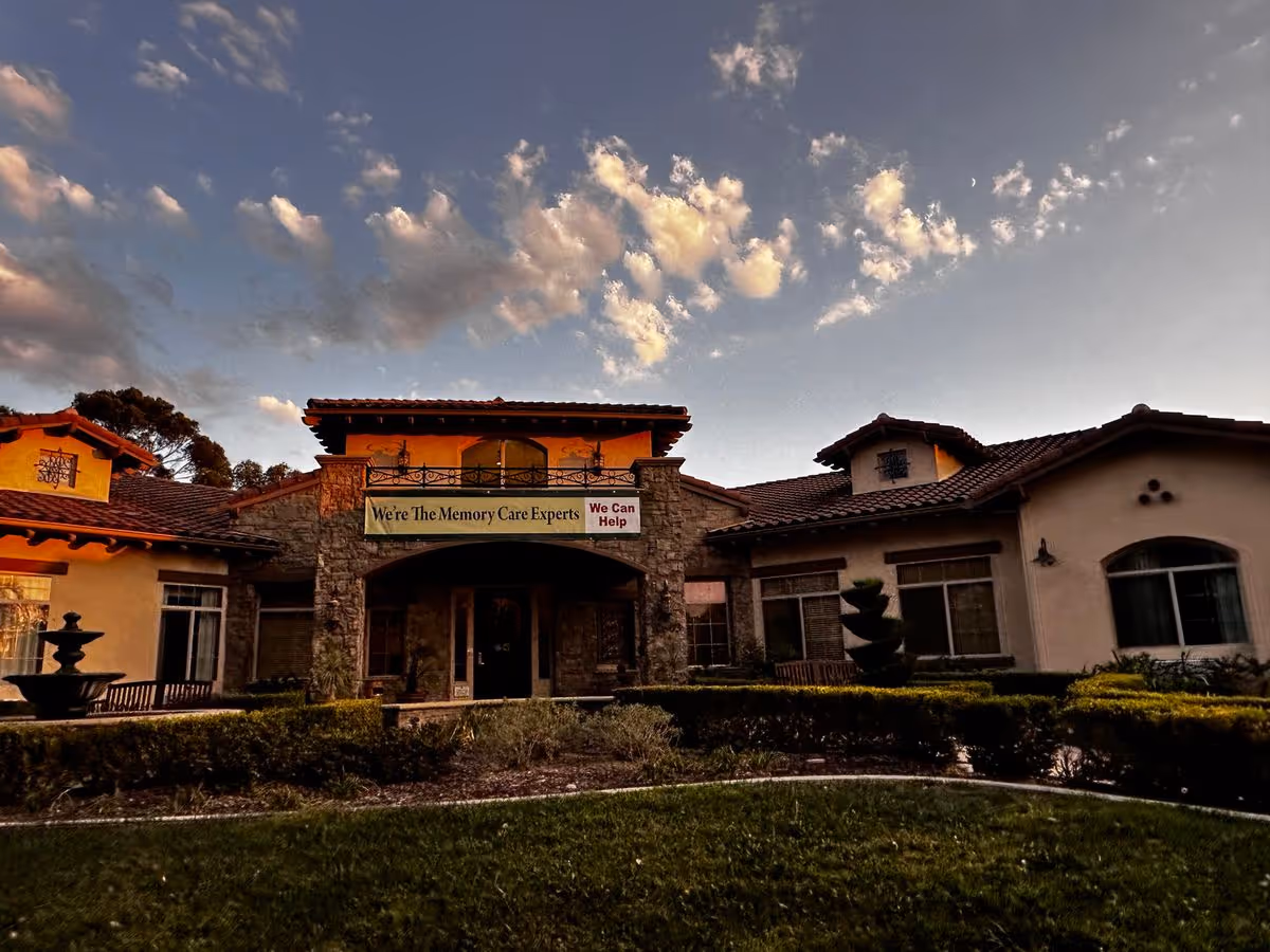 The front exterior of a Mediterranean-style senior care building with a banner reading "We're The Memory Care Experts" beneath a dramatic cloudy sky.