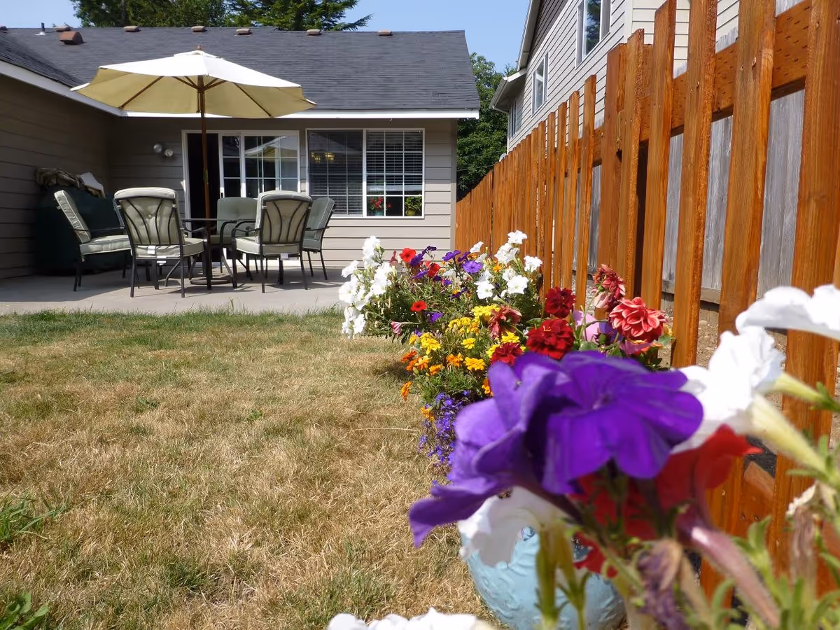 Outdoor patio area with a table, six cushioned chairs, and a large white umbrella. There is a wooden fence on the right side and a row of colorful flowers in the foreground. The patio is adjacent to a beige house with a window and a door.