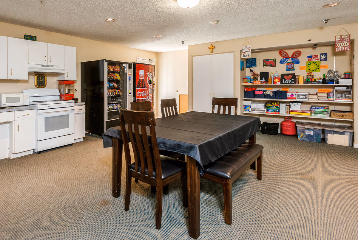 A communal room with a kitchen area featuring a white stove, microwave, and cabinets. There is a vending machine next to the kitchen. In the center, a dark wooden table with a black tablecloth is surrounded by four chairs and a bench. On the right wall, shelves hold various games, books, and colorful decorations. The room has beige walls and carpeted flooring.