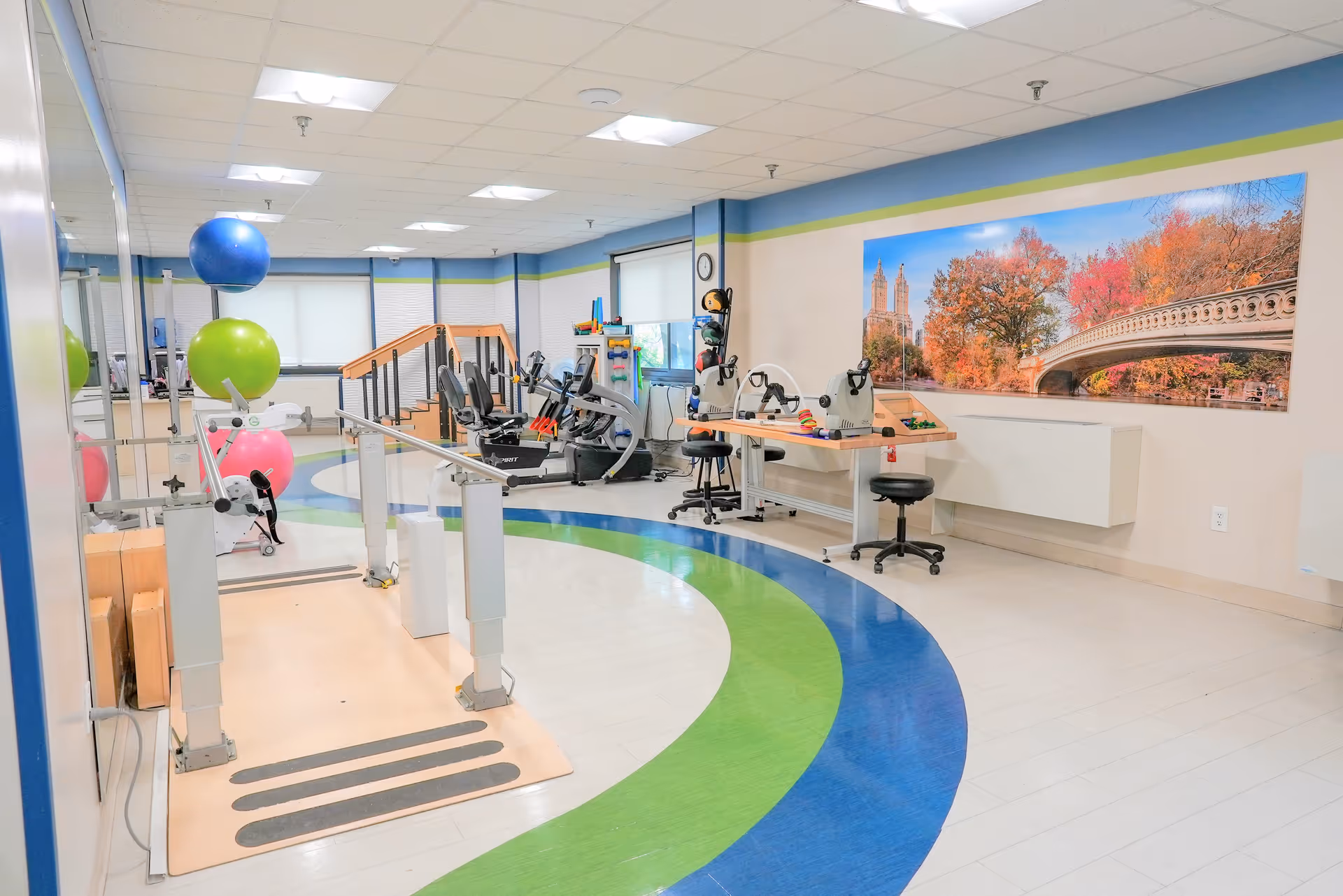 Bright rehabilitation gym with parallel bars, exercise bikes, therapy balls and workstations beneath a large wall mural.