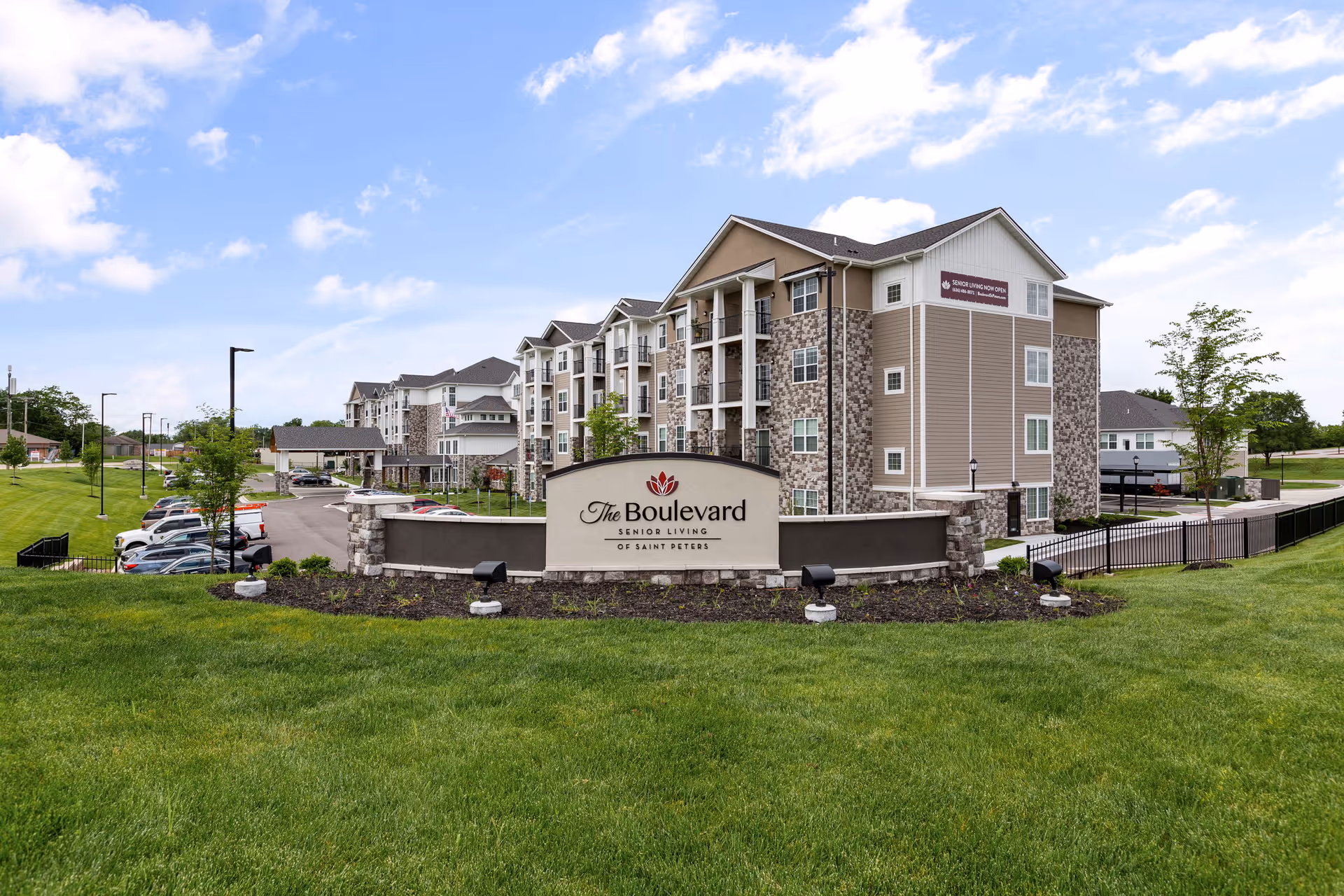 Front exterior of The Boulevard Senior Living of Saint Peters with its entrance sign, landscaped lawn, parking area and multi-story building.