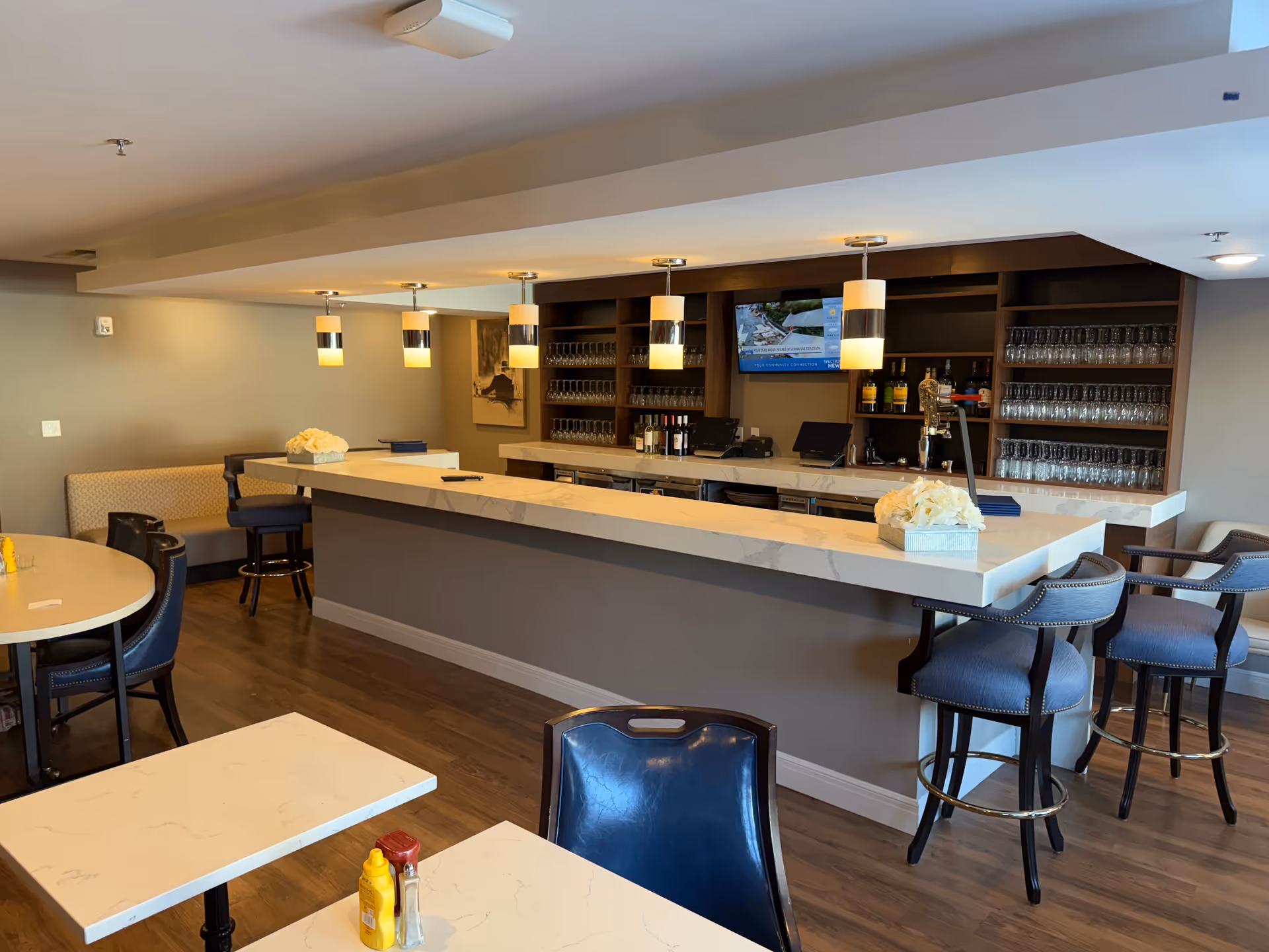 Interior view of a senior living facility's bar and dining area with a long marble countertop bar, bar stools, hanging pendant lights, shelves with glasses and bottles, and tables with chairs. The room has wooden flooring and neutral-colored walls.