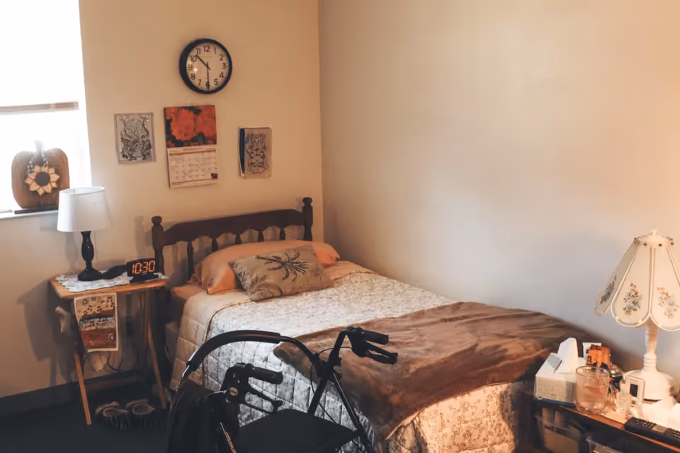 A cozy bedroom with a single bed covered in a floral bedspread and a brown blanket. There is a wooden headboard, a decorative pillow, and a walker in front of the bed. A small wooden side table holds a lamp, an alarm clock showing 10:30, and a decorative cloth. On the wall above the bed are a clock, a calendar, and some artwork. Another table with a floral lamp, tissues, and various small items is beside the bed. A window with a pumpkin decoration is visible on the left side.