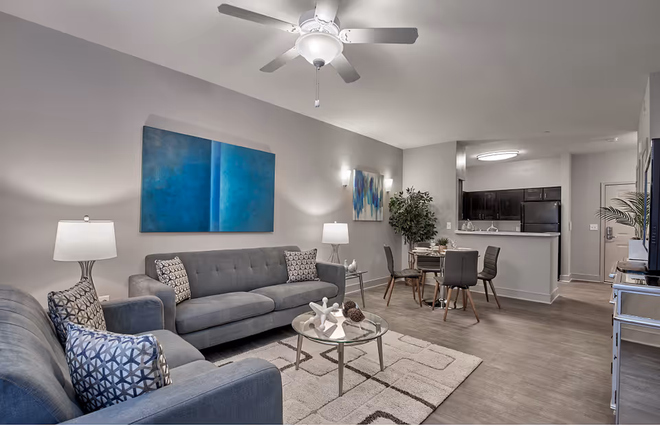Open-plan living room and dining area with gray sofas, a glass coffee table, dining table, and kitchen in the background.