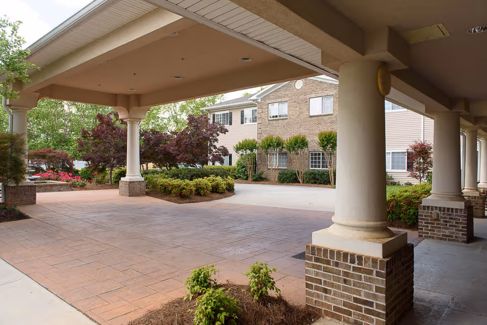 Covered entrance area with large columns and brick bases leading to a driveway surrounded by landscaped bushes and trees, with a multi-story building in the background.