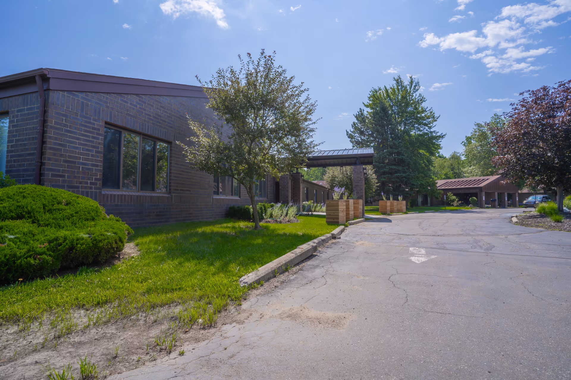 Front exterior of a single-story brick senior care facility with a driveway, covered entrance, trees, and landscaping.