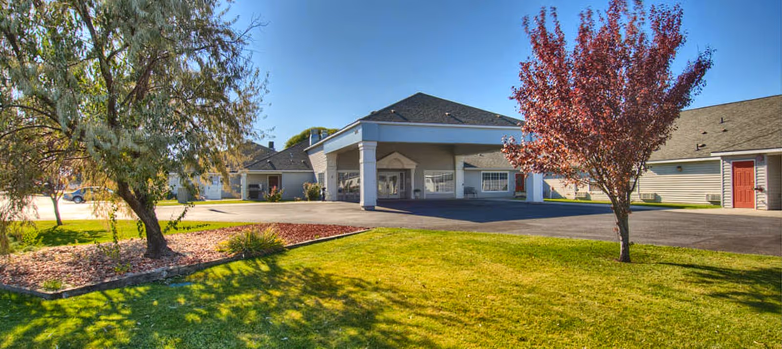 Exterior view of Dorian Place Assisted Living facility showing a single-story building with a covered entrance, surrounded by a well-maintained lawn, trees, and a clear blue sky.
