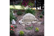 A memorial garden area with a large engraved stone surrounded by smaller rocks and various flowering plants. There is a small statue of a child on the left side of the stone. Trees and greenery are visible in the background.