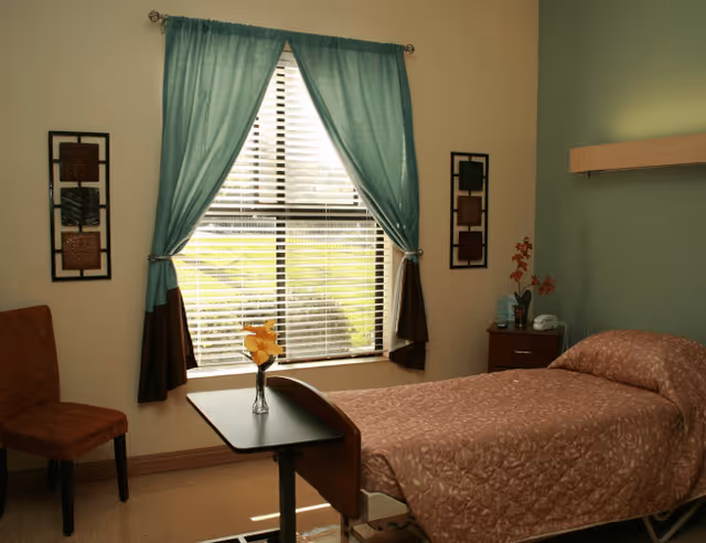 A single bed with a brown patterned bedspread in a room with a window covered by teal and brown curtains. There is a small table with a vase of yellow flowers in front of the bed, a brown chair to the left, and a nightstand with a phone and a small plant to the right. The walls are painted in light beige and green tones.