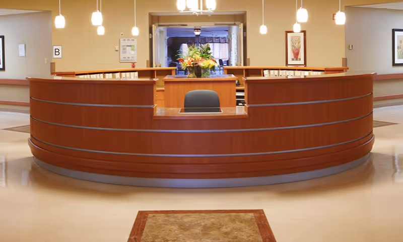 A large curved wooden reception desk with a chair behind it, a flower arrangement on the desk, and pendant lights hanging from the ceiling in a well-lit interior space.