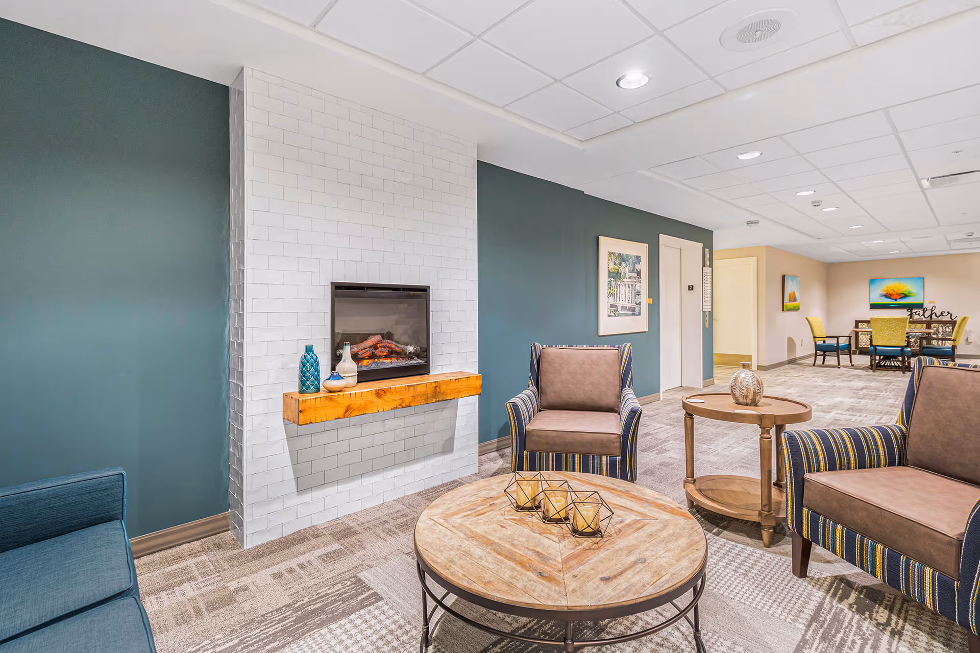 Well-lit common seating area with armchairs, round coffee tables, and a wall-mounted fireplace in a senior living facility.