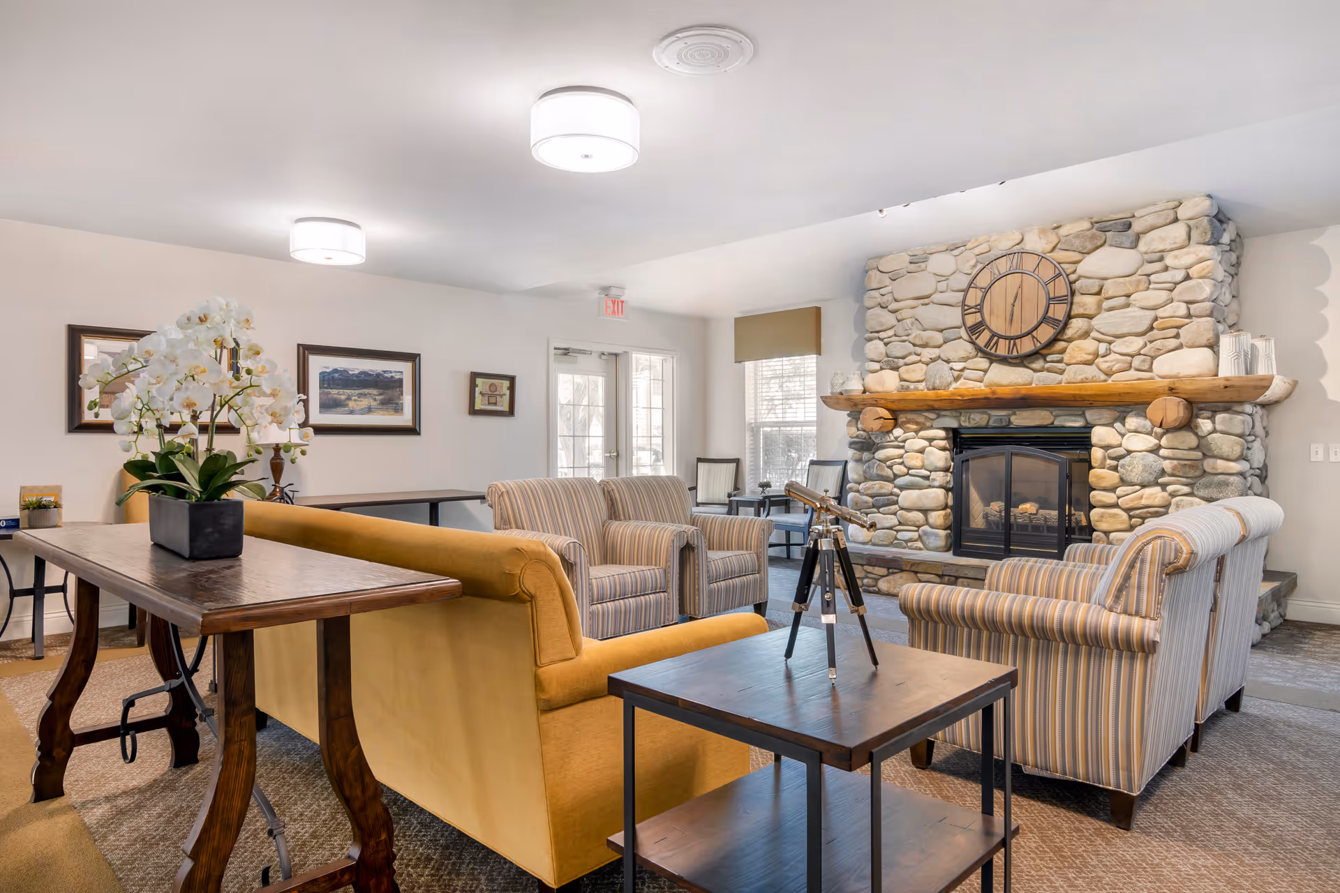 A cozy living room area with a stone fireplace featuring a large wooden clock above it. The room has striped armchairs and a yellow sofa arranged around a wooden coffee table with a telescope on it. There is a wooden console table with a potted white orchid, framed pictures on the wall, and a door with windows letting in natural light.