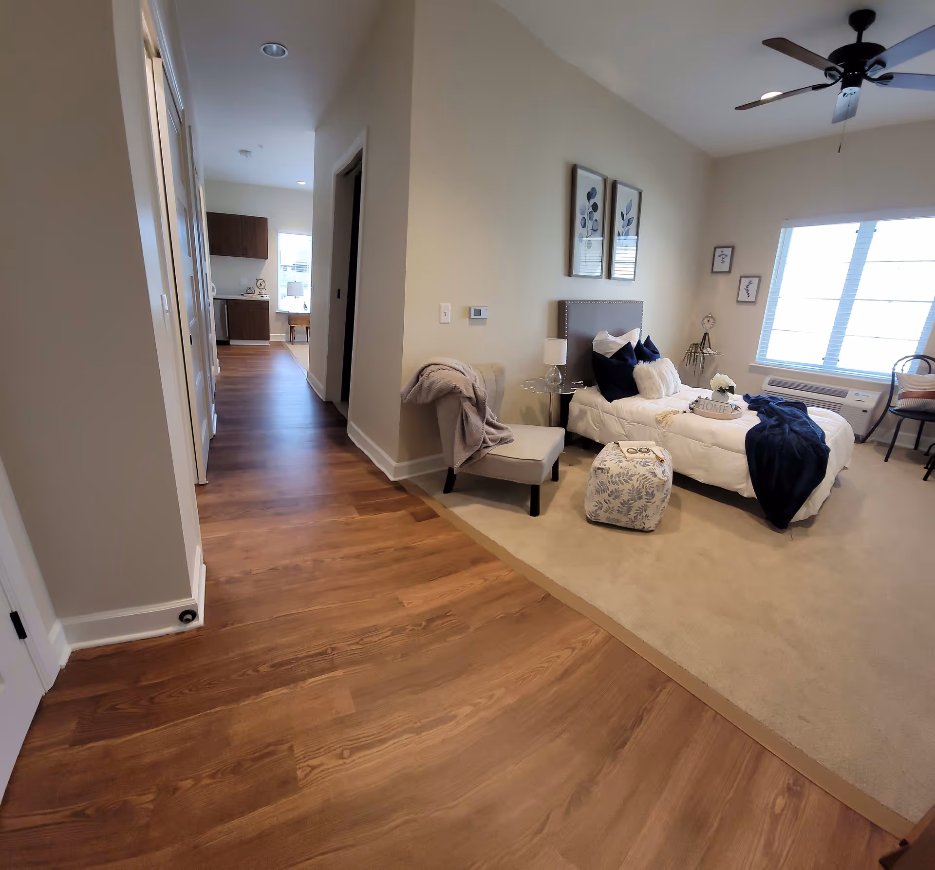 A bright and cozy bedroom in an assisted living facility with a single bed adorned with white and navy blue pillows and a navy blue throw blanket. The room features a beige carpet, a small upholstered chair with a blanket draped over it, a patterned ottoman, and framed botanical artwork on the wall. A ceiling fan is visible above, and a large window lets in natural light. The room opens to a hallway with wood flooring leading to a kitchen area in the background.