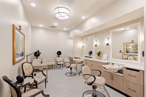 Interior view of a salon area in a senior living facility with beige walls and light wood flooring. The room features salon chairs in front of large mirrors mounted on a counter with cabinets below. There are additional chairs and a small table along the opposite wall, with framed artwork and modern light fixtures on the ceiling and walls.