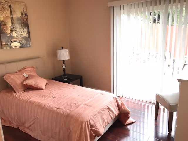 A bedroom with a single bed covered in a pink bedspread and matching pillows. Next to the bed is a black nightstand with a lamp on it. There is a large window with vertical blinds letting in natural light. A chair is positioned near the window, and a framed picture of the Eiffel Tower hangs on the wall above the bed.