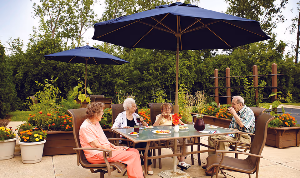 Four elderly people sitting around a glass outdoor table under large blue umbrellas, enjoying drinks and snacks in a garden area with green trees and colorful flowers in planter boxes.