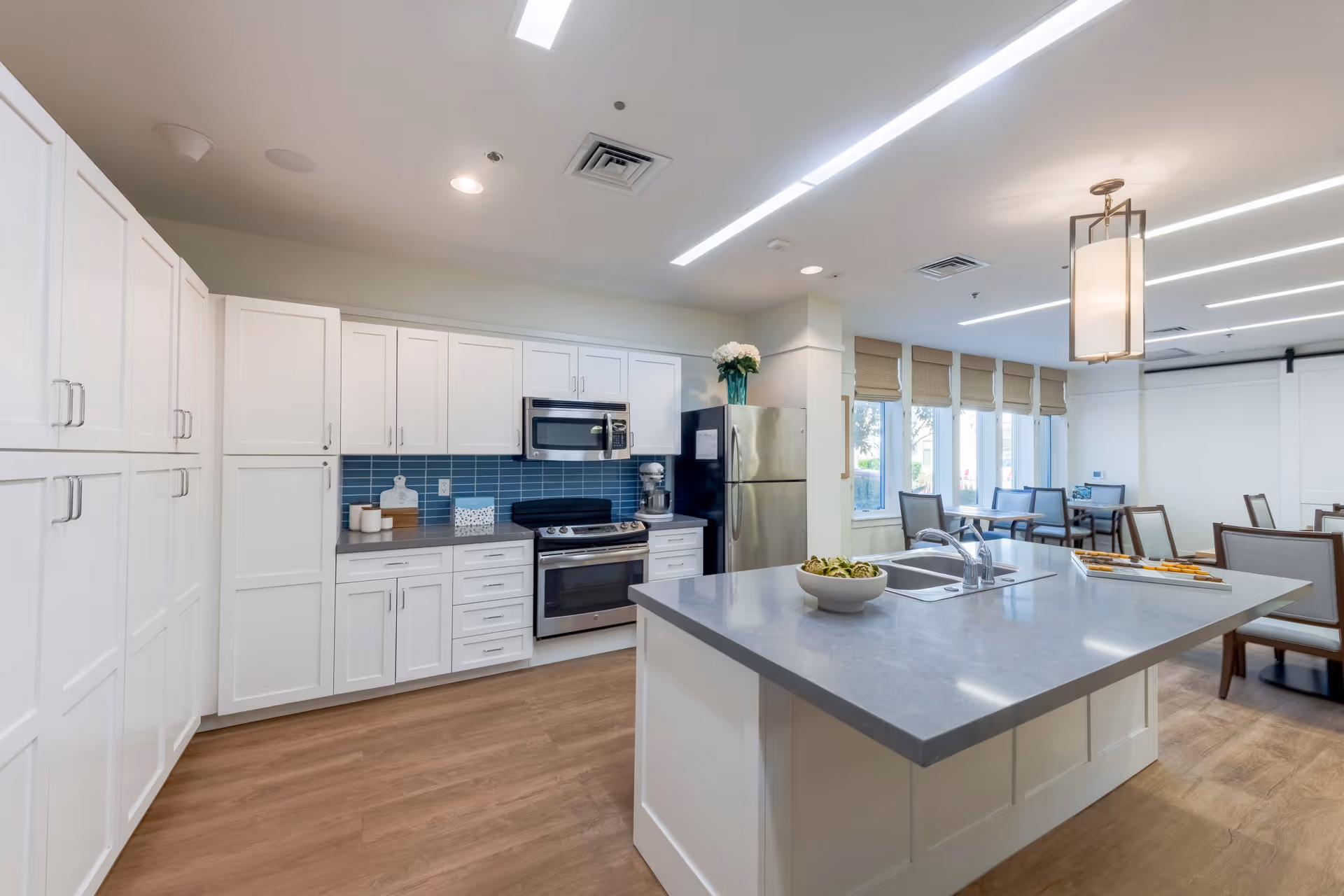 Modern kitchen area with white cabinetry, stainless steel appliances including a refrigerator, oven, and microwave. A large island with a sink and a bowl of food is in the center. In the background, there is a dining area with tables and chairs next to windows with beige blinds.