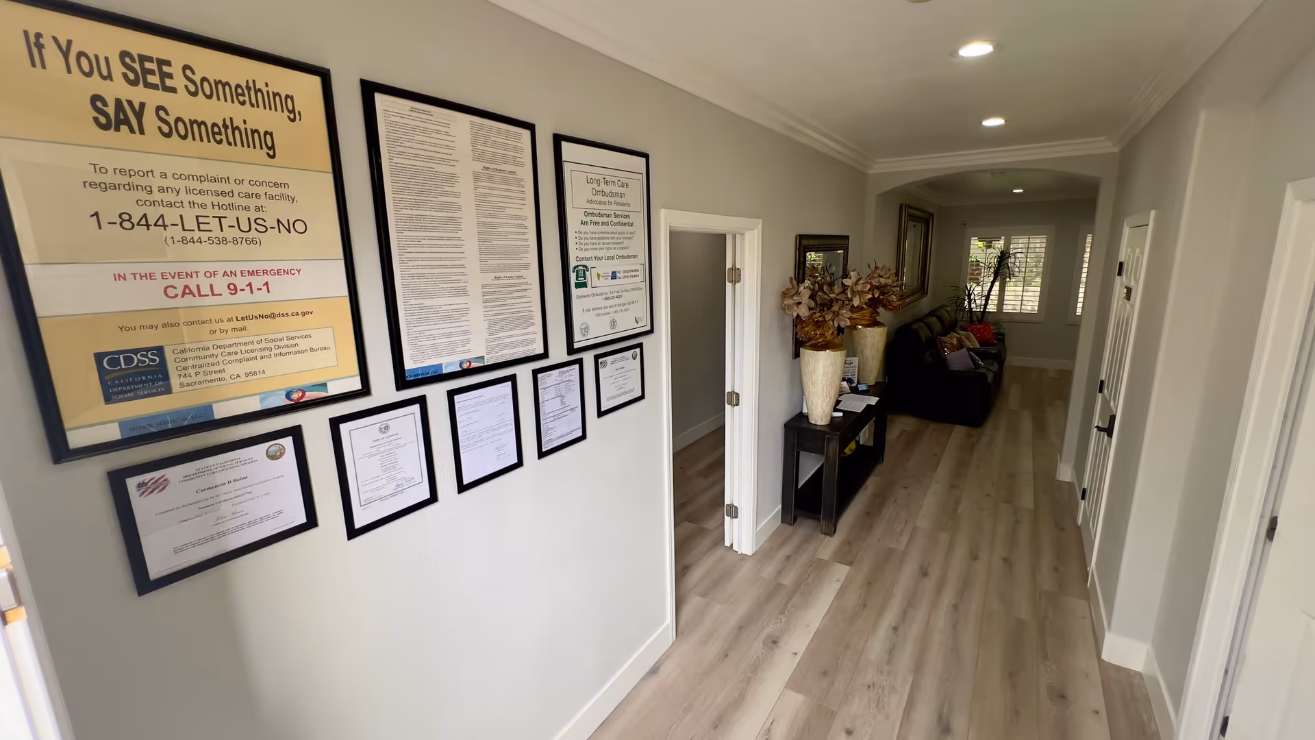 A hallway in a senior care facility with light wood flooring and light gray walls. On the left wall, there are multiple framed notices and certificates, including a large sign that says 'If You SEE Something, SAY Something' with emergency contact information. On the right side, there is a small black table with two large vases holding dried flowers, a mirror above the table, and a dark-colored couch with pillows near a window with white shutters. Recessed ceiling lights illuminate the space.