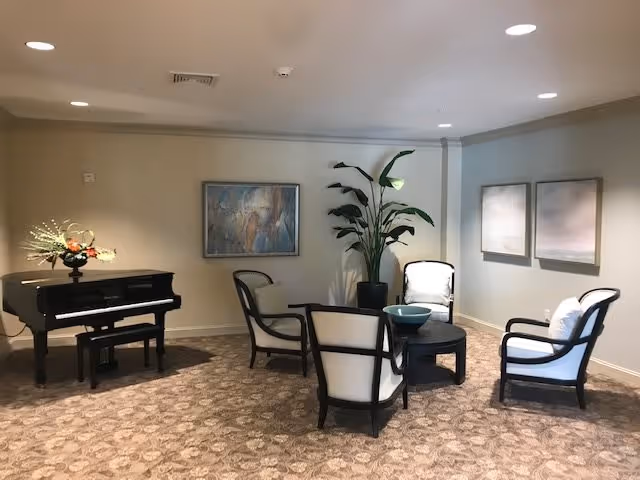 A cozy sitting area with four cushioned armchairs arranged around a round black coffee table with a decorative bowl on it. To the left, there is a black grand piano with a floral arrangement on top. The walls are adorned with three framed artworks, and a tall potted plant is placed in the corner. The room has a patterned carpet and recessed ceiling lights.