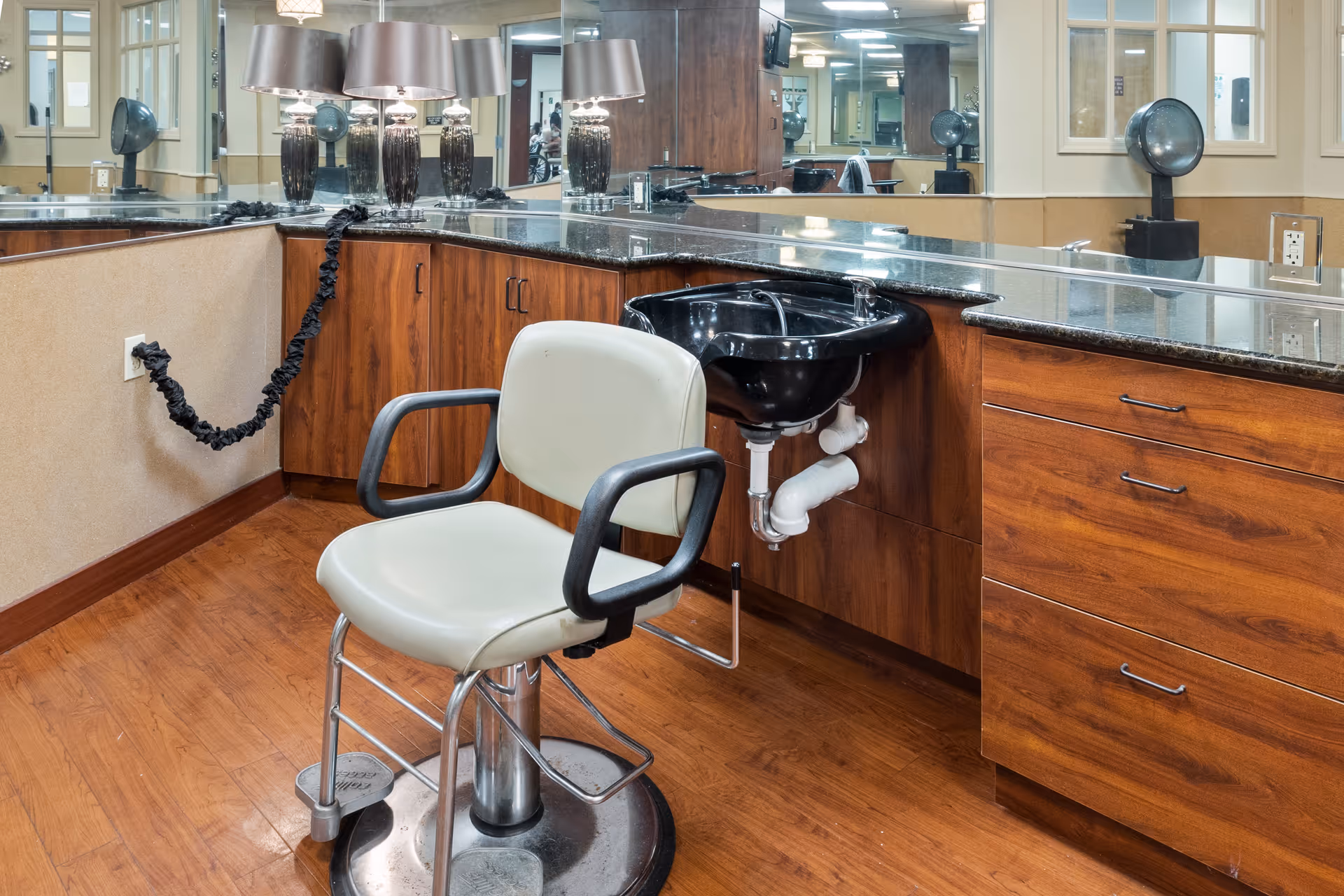 Interior view of a salon area in a senior living facility featuring a beige salon chair with black armrests in front of a black hair washing sink. The countertop is dark granite with wooden cabinets below, and a large mirror spans the wall behind the counter. Two decorative lamps are placed on the countertop, and the floor is wood.