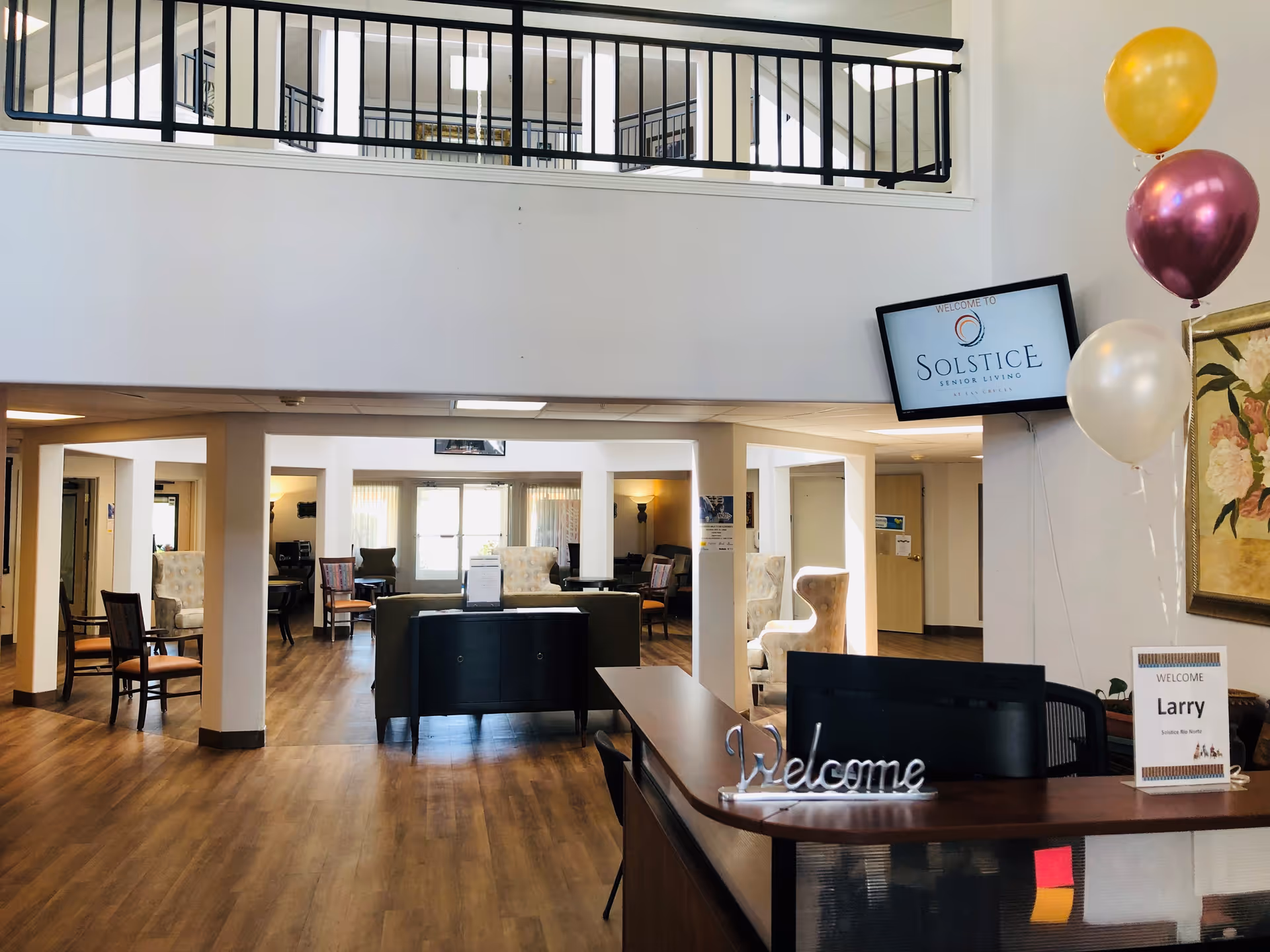 Reception area of Solstice Senior Living at Las Cruces featuring a wooden front desk with a 'Welcome' sign and a name card for Larry. Behind the desk, there is a seating area with chairs and sofas on a wooden floor. A TV mounted on the wall displays the Solstice Senior Living logo. Three balloons in gold, purple, and white are tied near the desk, and a floral painting hangs on the wall.