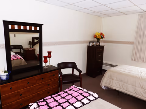 A furnished bedroom with two beds, a wooden dresser with mirror, a chair, and a chest of drawers with flowers on top.