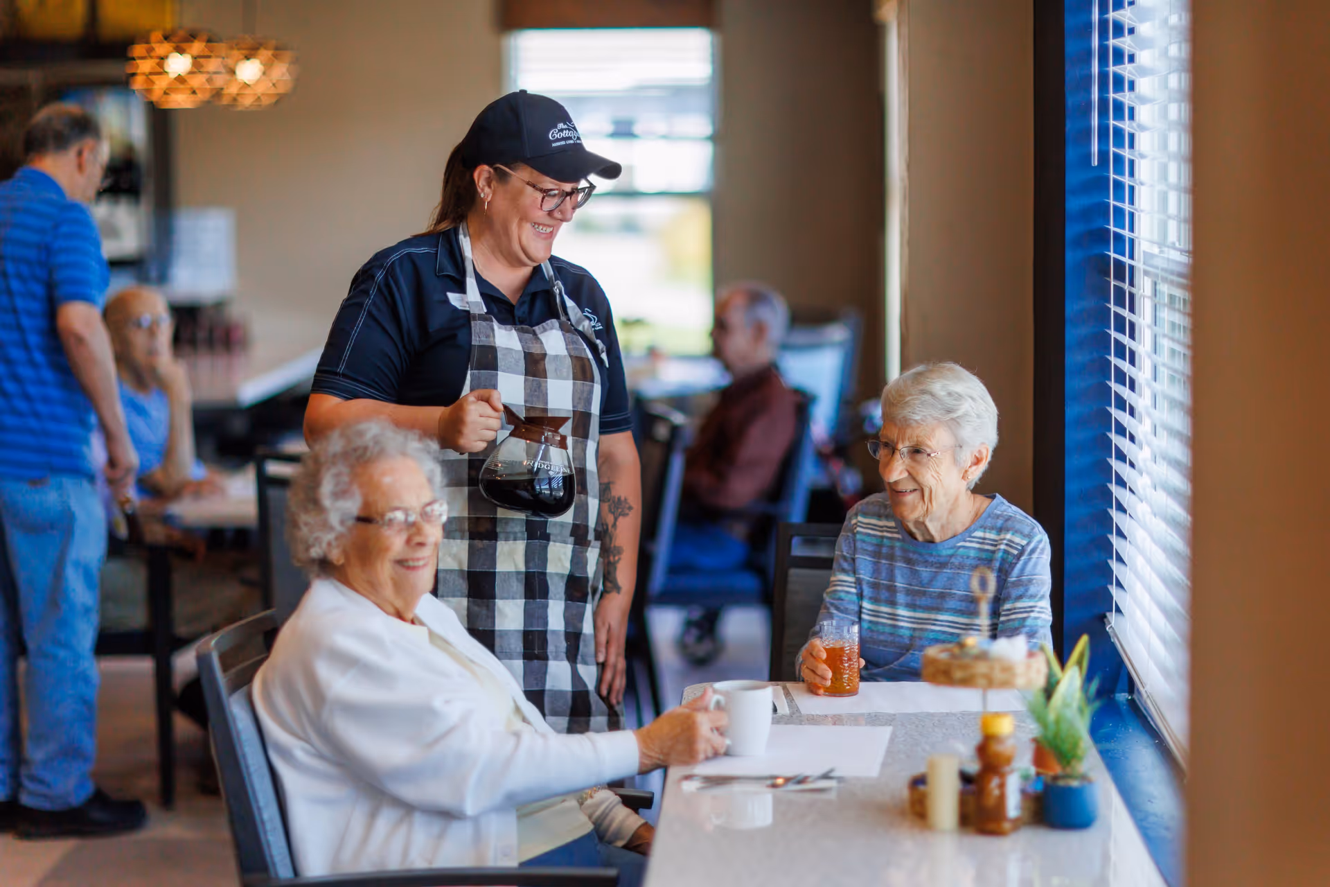 A smiling staff member wearing a black cap and checkered apron is pouring coffee for two elderly women seated at a table in a bright dining area. The women are enjoying drinks and conversation near a window with blinds. Other seniors are visible in the background sitting at tables.