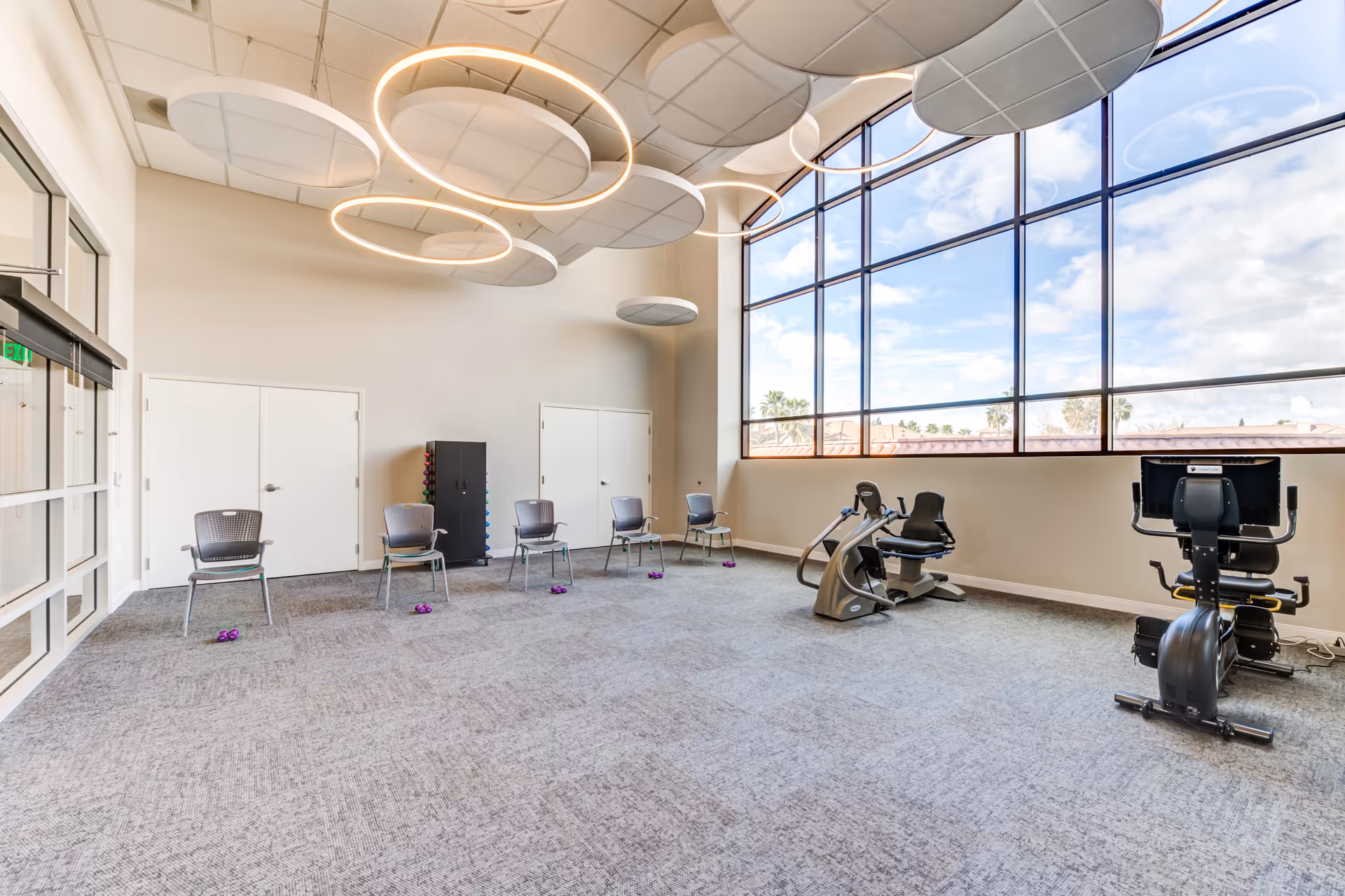 A spacious exercise room with large windows letting in natural light. The room has several chairs arranged in a row with small purple weights on the floor in front of each chair. There are two recumbent exercise bikes near the windows. The ceiling features modern circular light fixtures and acoustic panels.