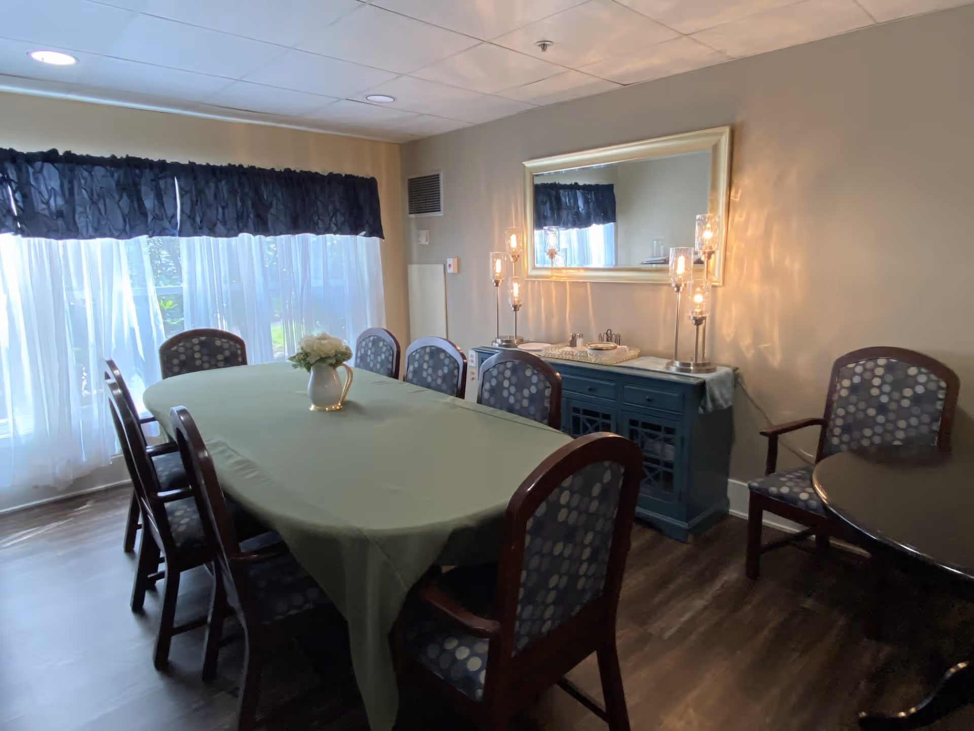 A dining room with a rectangular table covered with a green tablecloth, surrounded by eight chairs with patterned upholstery. A white vase with flowers is placed in the center of the table. Behind the table, there is a blue sideboard with decorative lamps and a large mirror hanging above it. Sheer white curtains with a dark valance cover the windows, allowing natural light to fill the room.