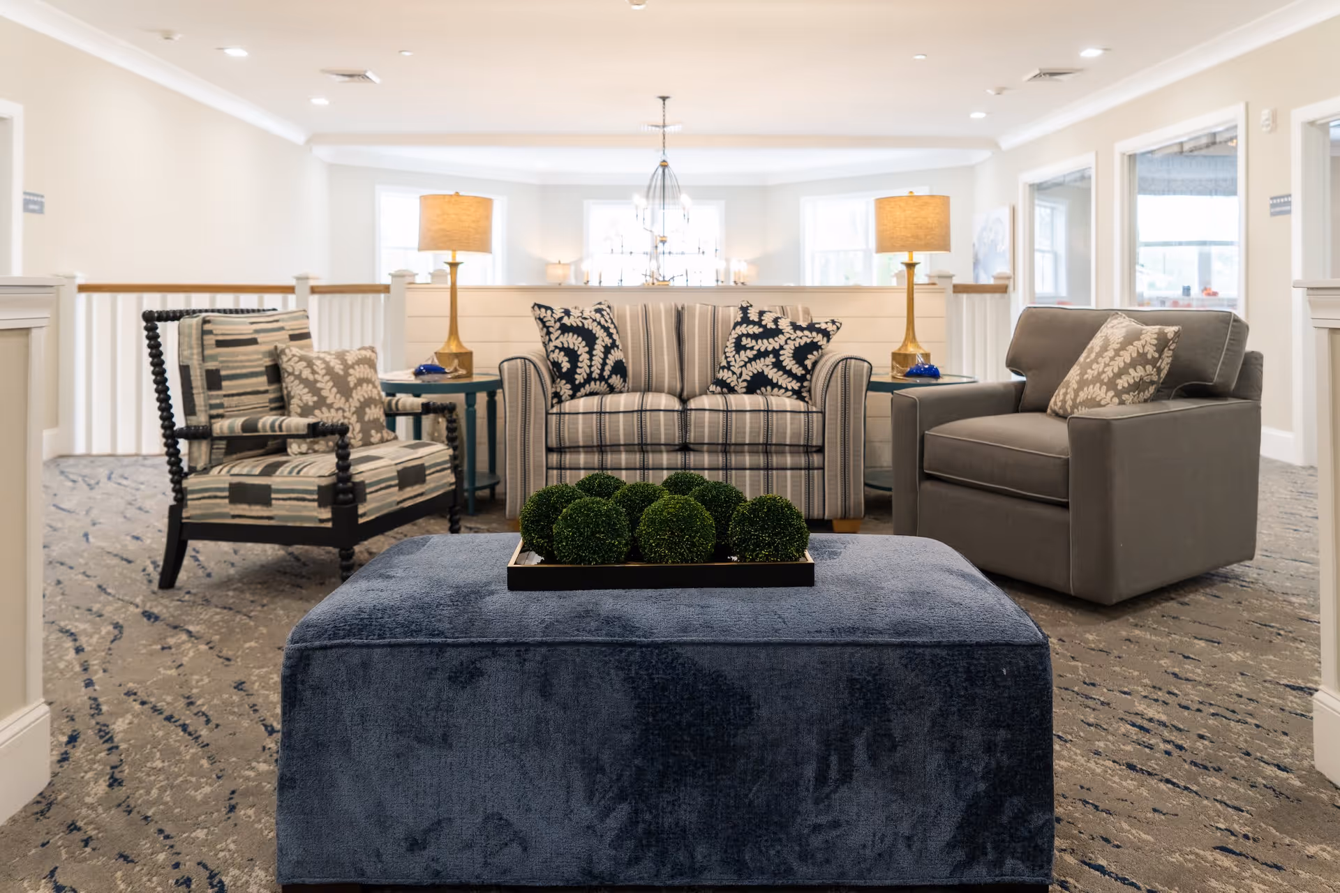 A cozy seating area in a senior living facility with a striped loveseat, a gray armchair, and a patterned armchair arranged around a large blue ottoman topped with a tray of decorative green spheres. Two brass table lamps with beige shades sit on side tables beside the loveseat. The space is well-lit with natural light from windows and has a carpeted floor with a subtle pattern.