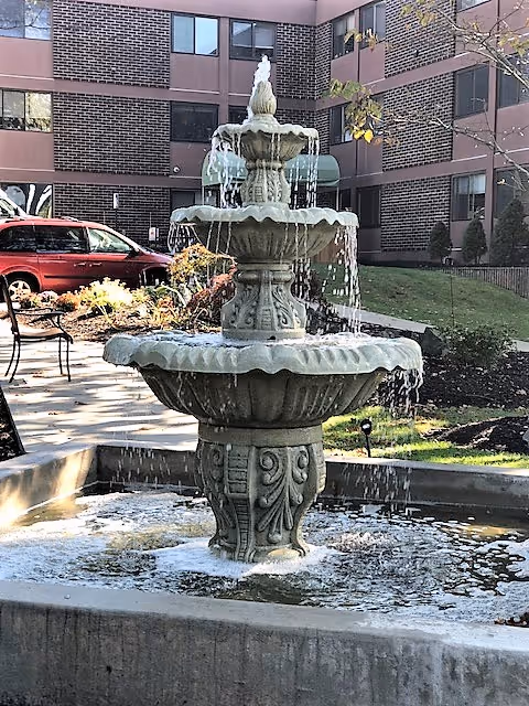 A multi-tiered stone water fountain with water flowing from the top tier down to the larger bottom basin, situated outdoors in front of a brick building with windows. There is a red vehicle parked to the left and some greenery including bushes and grass around the fountain.
