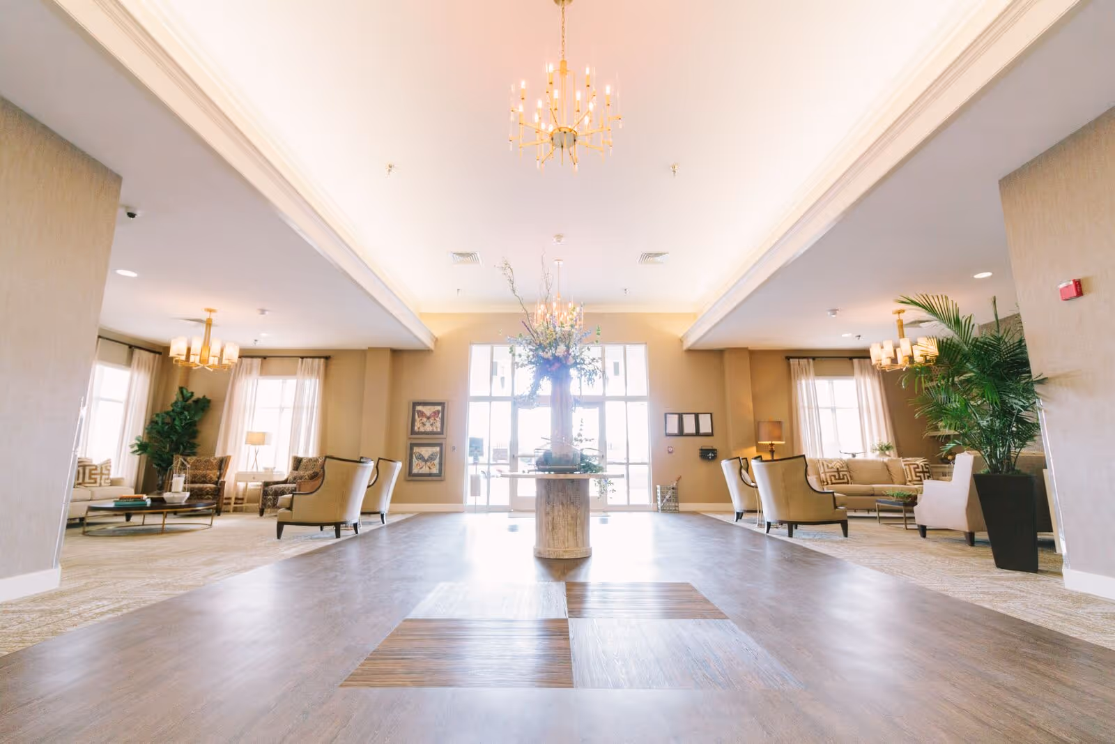 A spacious and well-lit common area with large windows letting in natural light. The room features comfortable seating arrangements with armchairs and sofas on either side, decorative plants, and a central table with a tall floral arrangement. Elegant chandeliers hang from the ceiling, and the floor is a combination of wood and carpet.