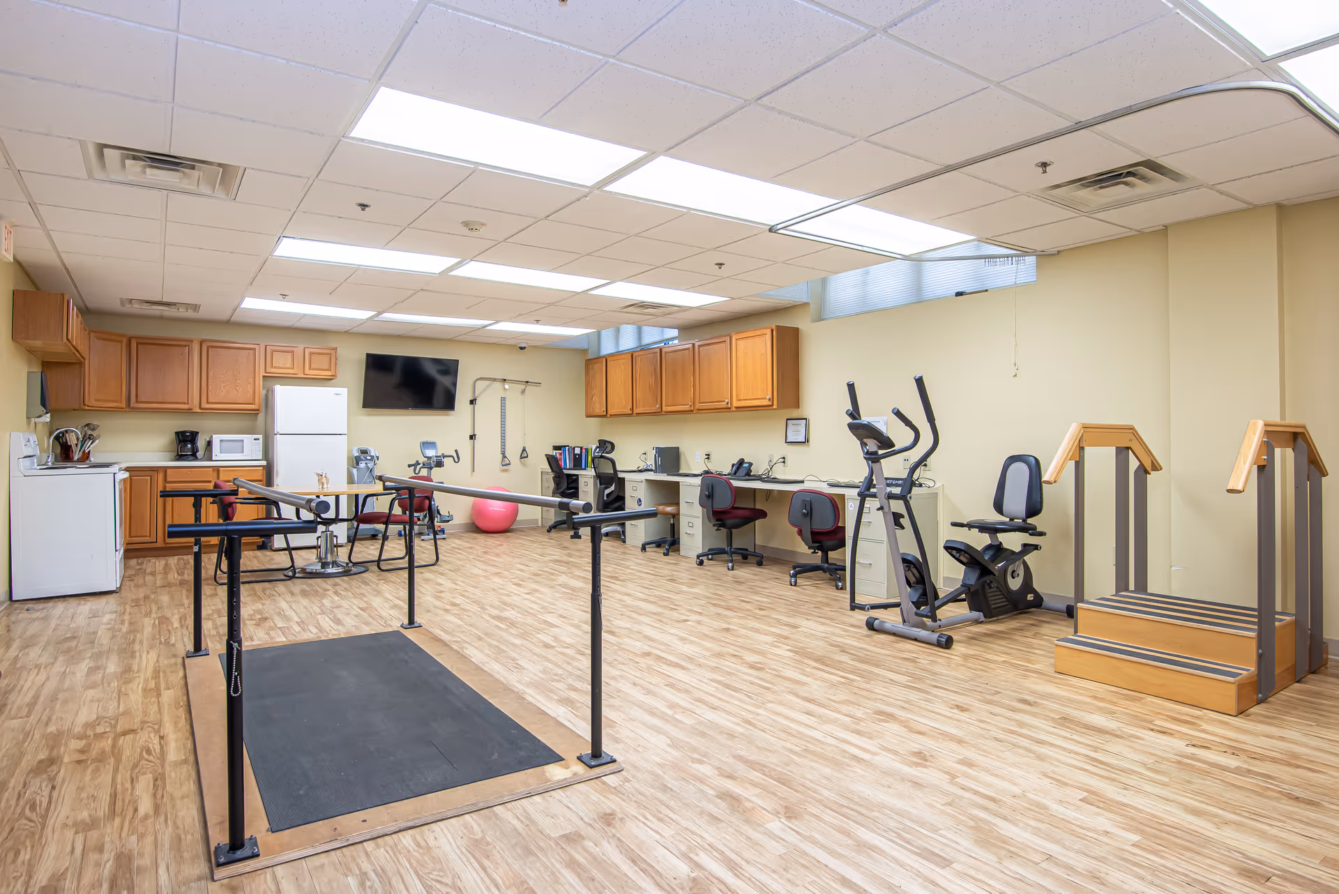 A spacious room with wooden flooring and beige walls featuring exercise equipment including parallel bars, a stationary bike, an elliptical machine, and a set of stairs for physical therapy. The room also has a kitchenette area with wooden cabinets, a refrigerator, microwave, and coffee maker. There are desks with office chairs and a wall-mounted TV.