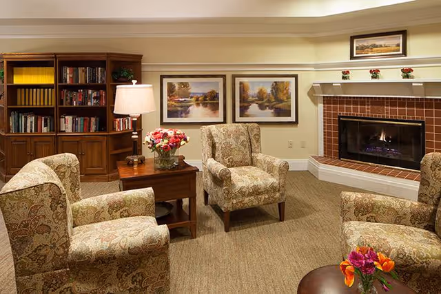 Cozy living room with four patterned armchairs arranged around a wooden side table with a lamp and a vase of flowers. A fireplace with a tiled surround and a mantle decorated with small flower pots is on the right. Behind the chairs, there is a wooden bookshelf filled with books and two framed landscape paintings on the wall.