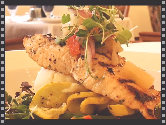 Grilled fish fillet atop mashed potatoes and sautéed vegetables, garnished with microgreens on a dining table.