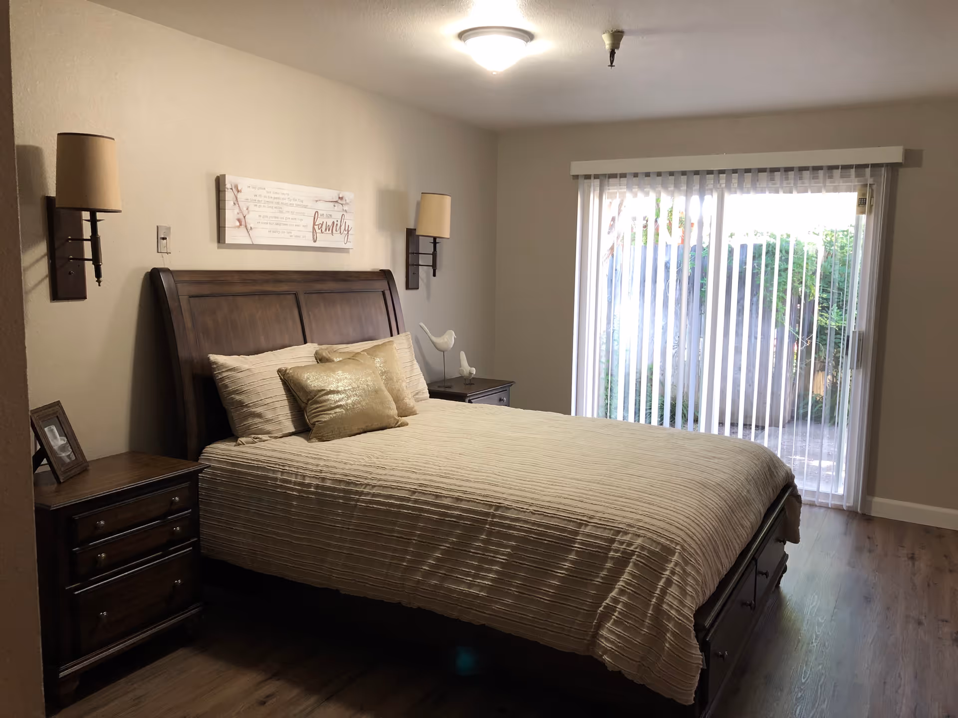 A bedroom with a wooden bed frame and beige bedding, two matching nightstands with lamps on either side, a framed picture on the left nightstand, a decorative bird figurine on the right nightstand, a wall decoration above the bed with the word 'family', and a large sliding glass door with vertical blinds leading to an outdoor area.