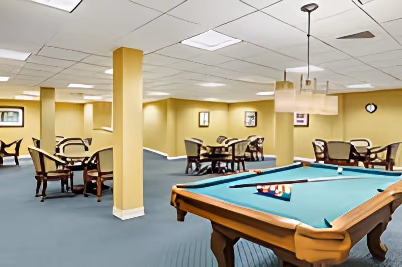 A spacious indoor recreational room with a pool table in the foreground and several tables with chairs arranged around the room. The walls are painted yellow and the floor is carpeted in blue. The ceiling has recessed lighting and a hanging light fixture above the pool table.