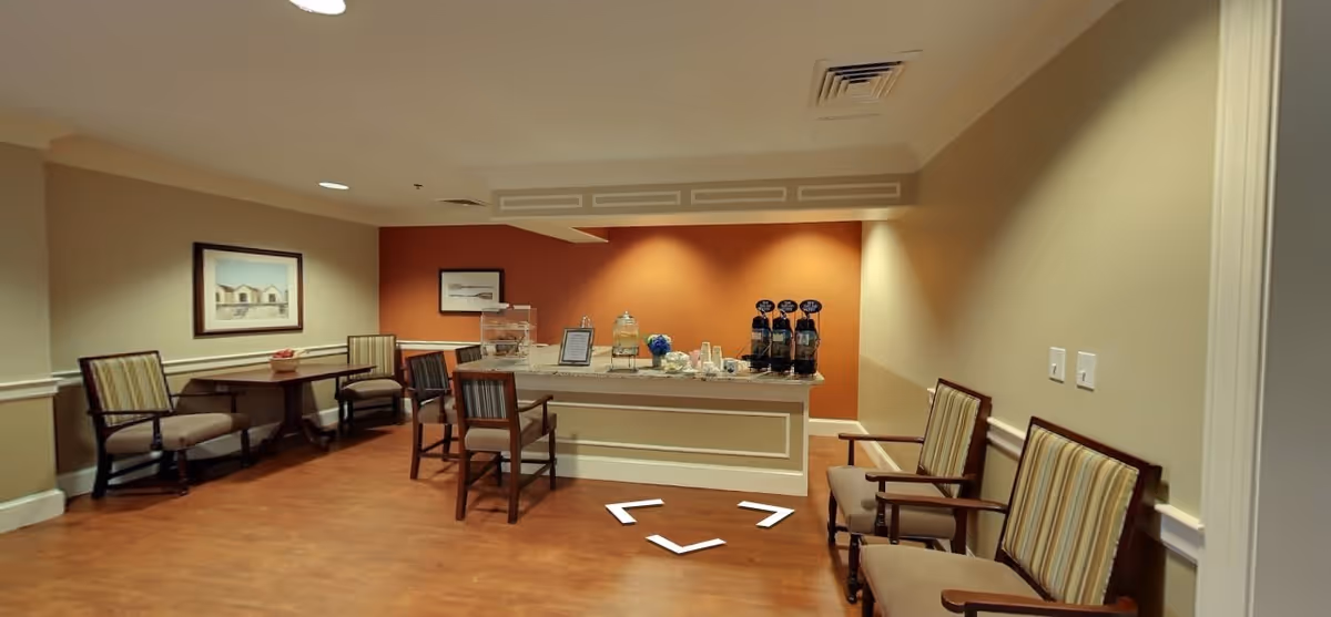 A small seating area with several wooden chairs with striped cushions arranged around small tables. A counter against a burnt orange accent wall holds beverage dispensers and cups. The floor is wooden, and the walls are painted beige with white trim. Two framed pictures hang on the walls.